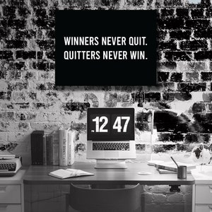 May include: A black and white photo of a home office with a brick wall, a desk, a laptop, a typewriter, books, and a plant. The laptop screen displays a digital clock showing 12:47. A motivational quote is displayed on the wall: "Winners never quit. Quitters never win."