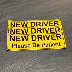 May include: Yellow sign with black text that reads "NEW DRIVER NEW DRIVER NEW DRIVER Please Be Patient".