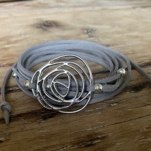 May include: A gray suede wrap bracelet with a silver metal rose design. The bracelet has multiple strands and is accented with small silver beads.