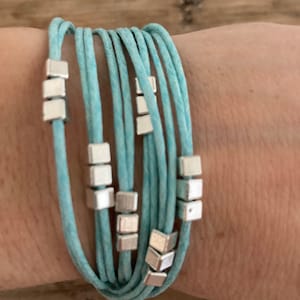 Può includere: Un braccialetto multifilo color turchese con perline quadrate argentate.