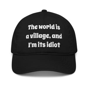 May include: A black baseball cap featuring white embroidered text on the front that reads, The world is a village, and I'm its idiot. The cap has a structured crown, a curved brim, and a button at the top centre of the fabric panels.