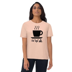Puede incluir: Una camiseta de manga corta en color melocotón claro con un gráfico negro de una taza de café humeante sobre un platillo. Debajo del platillo, hay tres símbolos negros distintos: una línea ondulada, una forma vertical y un triángulo.