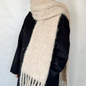 May include: A beige, fringed scarf is draped around a person's neck and shoulders. The scarf is made of a soft, fuzzy material. The person is wearing a black coat. The scarf's fringe hangs down.