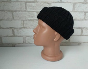 May include: A black ribbed knit beanie hat. The hat is folded at the brim and sits on a mannequin head. The hat is made of a thick, dark material and has a classic, simple design. The background is a white surface and a white brick wall.