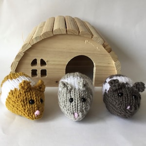 May include: Three knitted hamster toys in front of a wooden hamster house. The hamsters are white and brown, white and grey, and white and yellow.