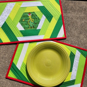 May include: Two green, white, and yellow quilted placemats with red trim. The top placemat features a green Grinch character in a Santa hat. The bottom placemat has a green plate on top.
