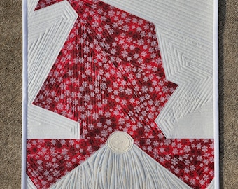 May include: A white and red quilted wall hanging with a geometric design. The design features a red fabric with a white snowflake pattern and a white fabric with a quilted pattern. The wall hanging is framed with a white border.