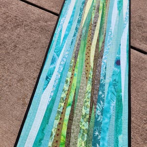 May include: A rectangular quilted table runner with a black border. The design features vertical stripes in shades of blue, green, and brown. The fabrics have various patterns, including floral and abstract designs.