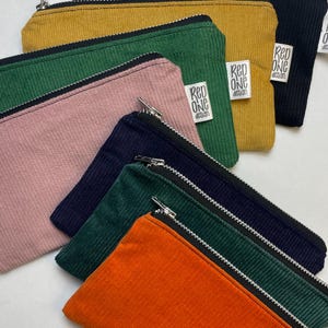 May include: A set of eight zippered pouches in various colors, including orange, green, pink, yellow, and blue. The pouches are made of corduroy fabric and have a black zipper. Each pouch has a white label with the text "Red One Design".