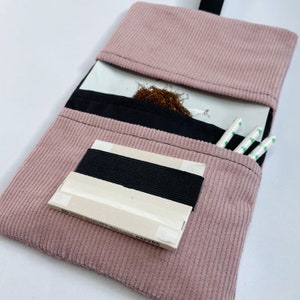 May include: A pink corduroy pouch with a black flap. The pouch has a white tray inside with tobacco and a pack of rolling papers. There are also three white cigarette filters with green tips.