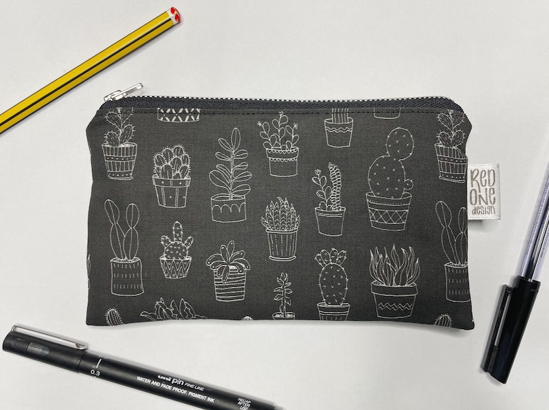 May include: A black zippered pouch with a white cactus and succulent print. The pouch has a white label with the text "Red One Design".