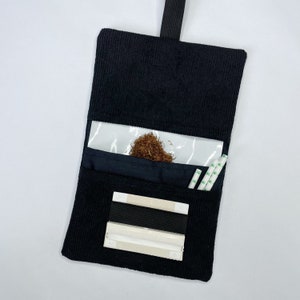 May include: Black corduroy pouch with a clear plastic window, a black elastic band, and a white rolling paper holder. The pouch is open and there is tobacco inside the window.