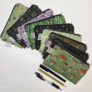 May include: A set of 10 zippered pouches with various nature-themed prints, including mushrooms, plants, and cacti. The pouches are made of fabric and have a green, black, and white color scheme. The pouches are arranged in a row, with the zippered ends facing the viewer. The pouches are all different sizes and shapes.
