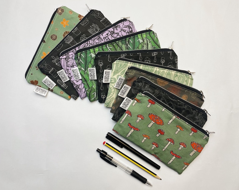 May include: A set of 10 zippered pouches with various nature-themed prints, including mushrooms, plants, and cacti. The pouches are made of fabric and have a green, black, and white color scheme. The pouches are arranged in a row, with the zippered ends facing the viewer. The pouches are all different sizes and shapes.