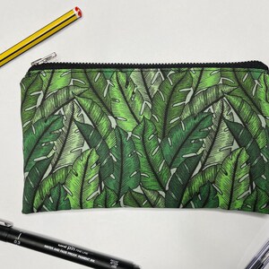 May include: A green zippered pouch with a tropical leaf pattern. The pouch has a black zipper and a white label that says "Red One Design".