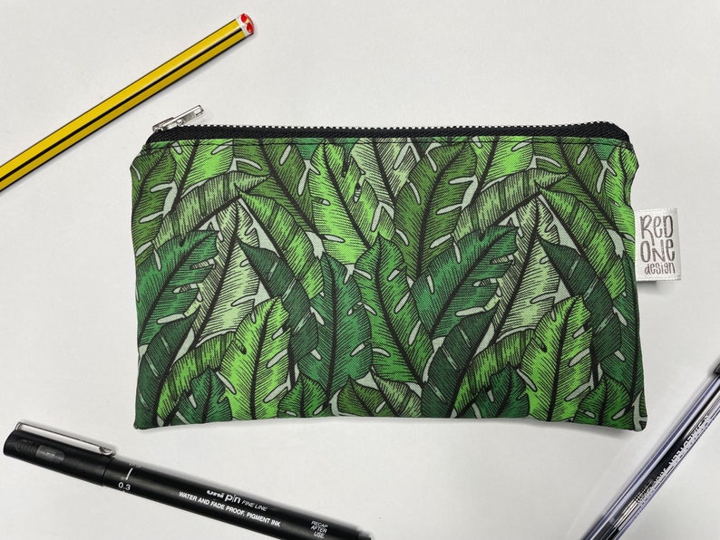 May include: A green zippered pouch with a tropical leaf pattern. The pouch has a black zipper and a white label that says "Red One Design".