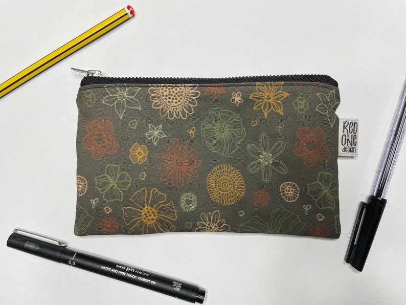 May include: A small zippered pouch with a floral print in shades of brown, yellow, and orange. The pouch has a black zipper and a small label that says "Red One Design".