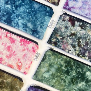 May include: A collection of eight colorful zippered pouches with tie-dye patterns. The pouches are made of fabric and have a black zipper. Each pouch has a small white label with the text "Red One Design" on it.