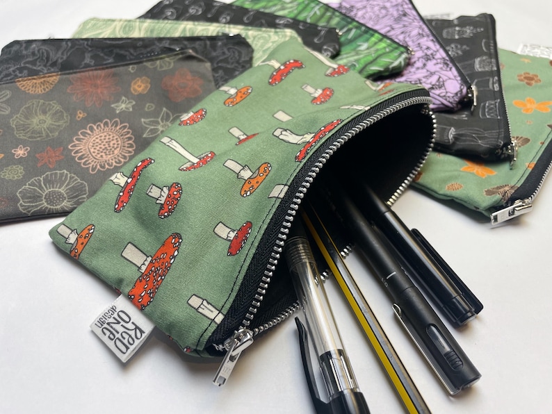 May include: A green zippered pouch with a mushroom print and the text "Red One" on the bottom. The pouch is filled with pens and pencils.