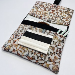 Può includere: Una pochette in tessuto con motivo bianco e marrone con una cerniera nera e un elastico nero. La pochette ha una tasca in plastica trasparente e uno slot per le cartine. La pochette è progettata per contenere cartine, tabacco e altri accessori per fumare.