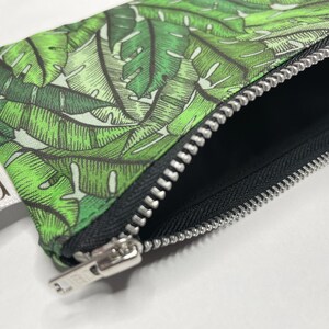 May include: A green and white zippered pouch with a tropical leaf print. The pouch has a silver zipper and a white label that reads "Red One Design".
