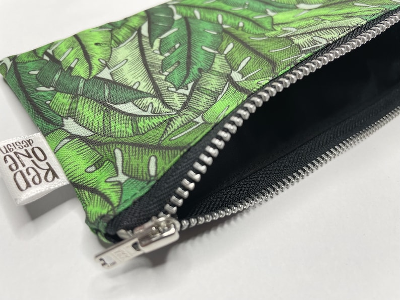 May include: A green and white zippered pouch with a tropical leaf print. The pouch has a silver zipper and a white label that reads "Red One Design".