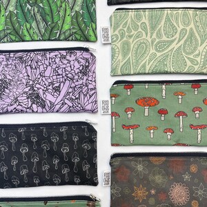 May include: Nine different zippered pouches with various patterns. The pouches are made of fabric and have a black zipper. The patterns include green leaves, paisley, mushrooms, flowers, and a black and white design. The pouches are all lined up in a row, with the top pouch being the largest and the bottom pouch being the smallest.