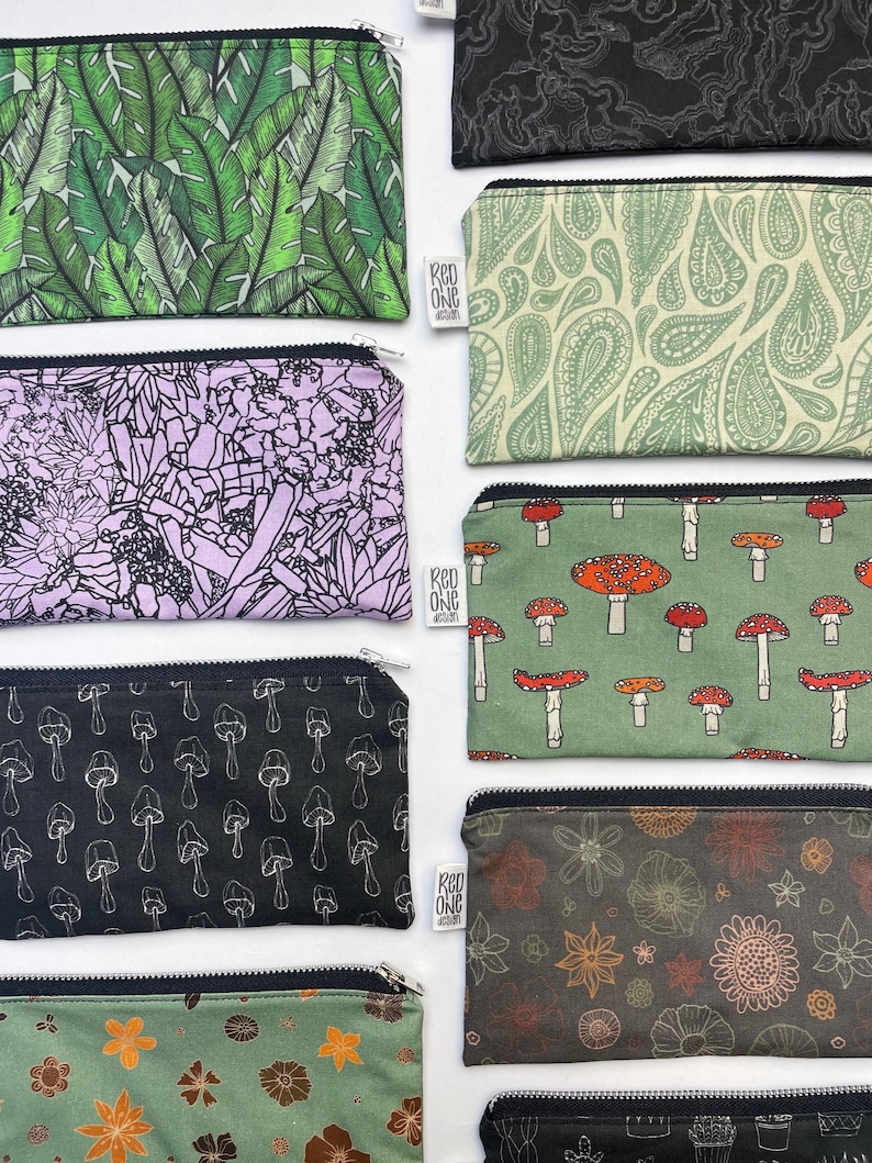 May include: Nine different zippered pouches with various patterns. The pouches are made of fabric and have a black zipper. The patterns include green leaves, paisley, mushrooms, flowers, and a black and white design. The pouches are all lined up in a row, with the top pouch being the largest and the bottom pouch being the smallest.