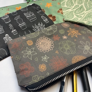 May include: Three zippered pouches with different floral and plant designs. The pouches are made of a soft, canvas-like material and have black zippers. The pouches are filled with pens and pencils.