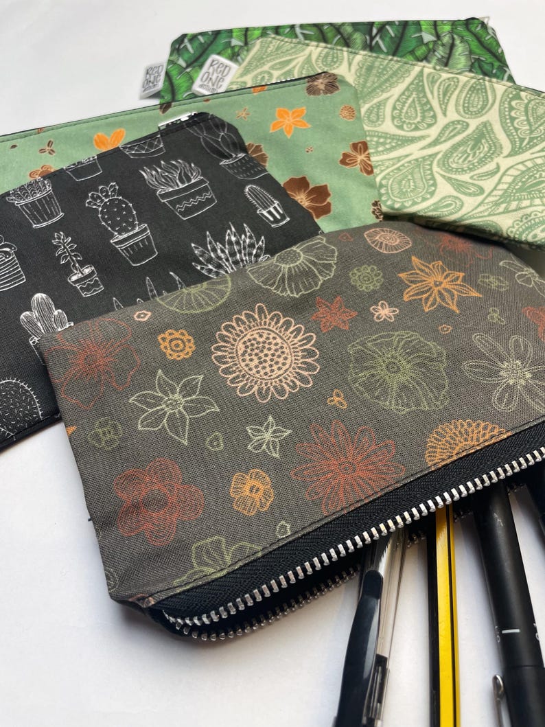 May include: Three zippered pouches with different floral and plant designs. The pouches are made of a soft, canvas-like material and have black zippers. The pouches are filled with pens and pencils.