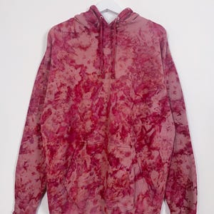 May include: A pink and red tie-dye hoodie hangs on a white hanger. The hoodie has a kangaroo pocket and a drawstring hood. The fabric displays a mottled pattern, with darker red areas contrasting with lighter pink hues.