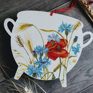 May include: A white ceramic cauldron with a floral design featuring red poppies and blue cornflowers. The cauldron has two handles and three legs. The background is a wooden surface.