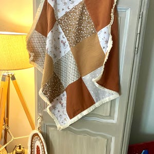 Peut inclure: Une couverture patchwork pour bébé composée de carrés alternés en tissu rouille uni, en tissu blanc à motif floral marron et en tissu blanc à motif céleste marron, le tout bordé d'une frange de couleur crème sur tout le pourtour.