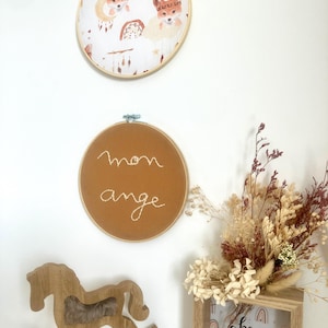 Puede incluir: Dos adornos de pared redondos con aros de madera. Uno presenta zorros y un diseño de atrapasueños, el otro tiene bordadas las palabras "mon ange". También se ven un caballito balancín de madera y una impresión enmarcada con el texto "be happy today".