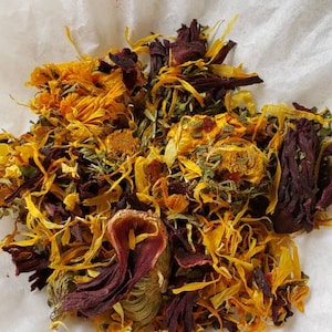 Op de afbeelding: Een close-up van een mengsel van gedroogde kruiden en bloemen, waaronder gele calendula-bloemblaadjes, rode hibiscus-bloemblaadjes en groene, bladerige kruiden. Het mengsel is gerangschikt op een wit oppervlak.