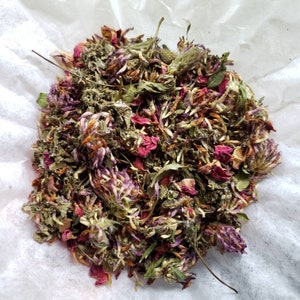 May include: A close-up shot of a pile of dried herbs and flowers. The mix includes purple clover-like flowers, red rose petals, and various green leaves. The blend is arranged on a white, textured surface.
