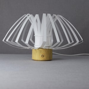 Puede incluir: Una lámpara de mesa blanca con un diseño geométrico único. La pantalla de la lámpara está hecha de tiras blancas delgadas que crean una forma de flor. La base es de madera y tiene un acabado marrón claro.
