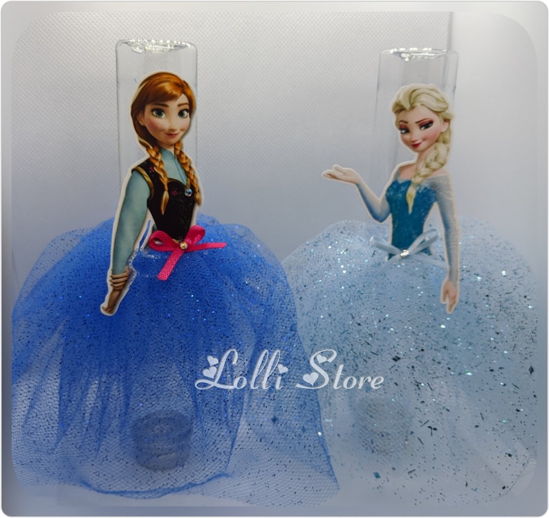 Puede incluir: Dos toppers de pastel de personajes de Frozen, uno con Anna y otro con Elsa. Ambos toppers llevan vestidos de tul azul con detalles de brillo. El texto "Lolli Store" es visible en la parte inferior central de la imagen.