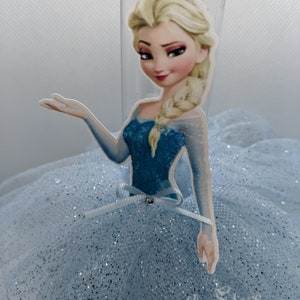 Puede incluir: Una falda de tul azul claro con detalles de purpurina y un recorte de cart&oacute;n de Elsa de Frozen. El recorte muestra a Elsa con un vestido azul y una trenza.