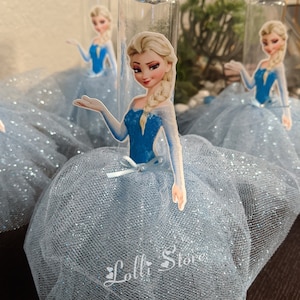 Puede incluir: Tres jarrones de vidrio transparente con tela de tul azul y purpurina. Cada jarr&oacute;n tiene una imagen impresa de Elsa de Frozen, con un vestido azul y una capa blanca. El texto "Lolli Store" est&aacute; impreso en la tela de tul.