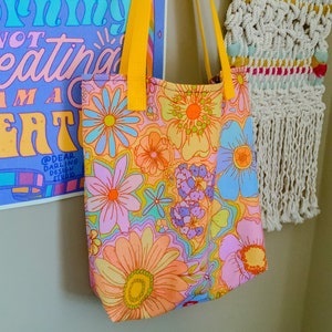 May include: A floral print tote bag with yellow straps. The bag features a colorful pattern of large flowers in shades of orange, pink, blue, and purple. The bag is hanging next to a poster with text and a macrame wall hanging.