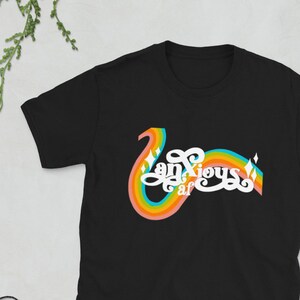 以下が含まれることがあります： レトロな虹のデザインで「Anxious af」の文字が入った黒いTシャツ。デザインは、虹のアーチが付いた、湾曲した様式化されたフォントが特徴です。シャツは柔らかい素材でできており、カジュアルウェアに適しています。