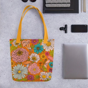 May include: Orange tote bag with a colorful floral and mushroom pattern. The bag has yellow handles and is surrounded by other items, including sunglasses, a notebook, pens, a phone, and a laptop.
