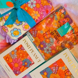 Puede incluir: Un conjunto de tres s&aacute;banas de cuna con estampado floral, una bata y una s&aacute;bana de cuna. Las s&aacute;banas son rosas con flores naranjas, amarillas, azules y moradas. La bata es rosa con flores naranjas, amarillas, azules y moradas. La s&aacute;bana de cuna es blanca con caritas sonrientes amarillas. El embalaje est&aacute; etiquetado con "Posh Peanut".