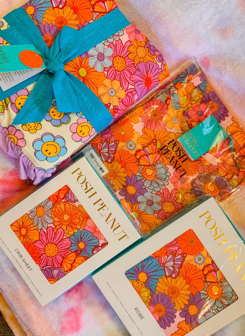Puede incluir: Un conjunto de tres s&aacute;banas de cuna con estampado floral, una bata y una s&aacute;bana de cuna. Las s&aacute;banas son rosas con flores naranjas, amarillas, azules y moradas. La bata es rosa con flores naranjas, amarillas, azules y moradas. La s&aacute;bana de cuna es blanca con caritas sonrientes amarillas. El embalaje est&aacute; etiquetado con "Posh Peanut".