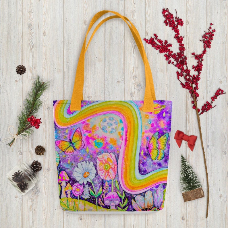 May include: A colorful tote bag with a whimsical design featuring a rainbow, butterflies, flowers, and mushrooms. The bag has yellow handles and a vibrant, psychedelic art style. The background is a mix of purple, pink, and blue.