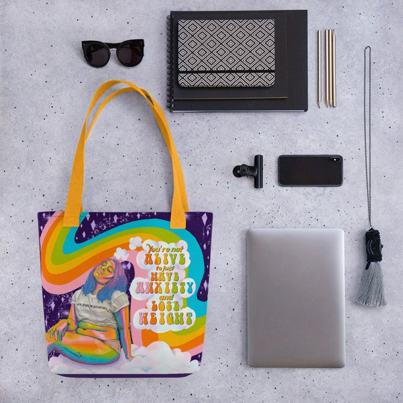 May include: A purple tote bag with yellow handles featuring a colorful illustration of a person sitting on a cloud with a rainbow and text that reads "You're not alive to just have anxiety and lose weight." Black sunglasses and a notebook are also visible.