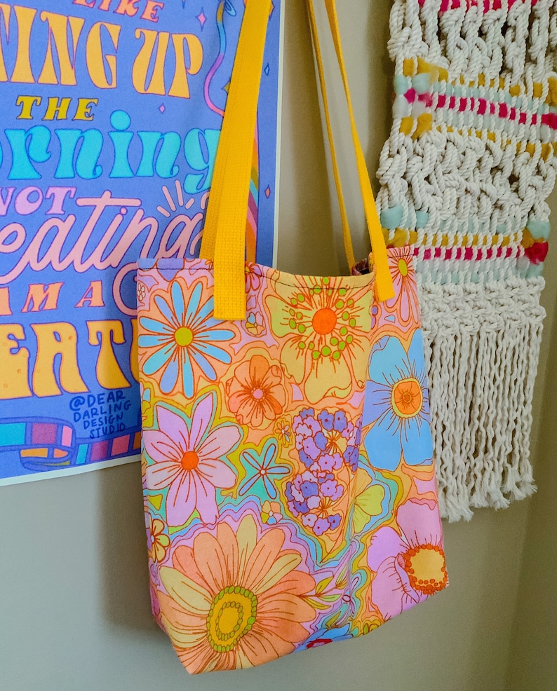 May include: A floral print tote bag with yellow straps. The bag features a colorful pattern of large flowers in shades of orange, pink, blue, and purple. The bag is hanging next to a poster with text and a macrame wall hanging.