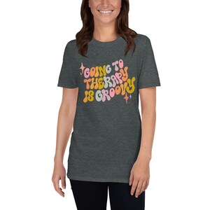 Puede incluir: Una camiseta gris oscuro jaspeada con un gr&aacute;fico de estilo retro que dice "Going to Therapy is Groovy". El gr&aacute;fico est&aacute; en tonos de rosa, naranja y amarillo.