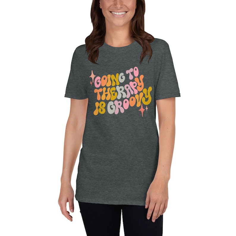 Puede incluir: Una camiseta gris oscuro jaspeada con un gr&aacute;fico de estilo retro que dice "Going to Therapy is Groovy". El gr&aacute;fico est&aacute; en tonos de rosa, naranja y amarillo.